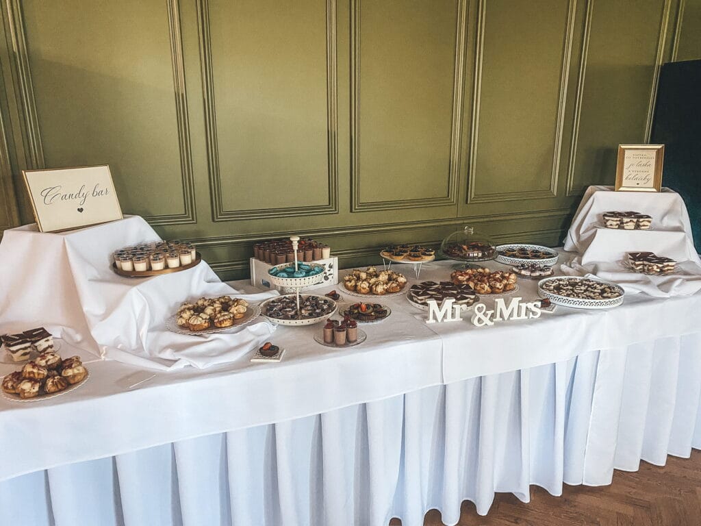 Svadobný candy bar s elegantným bielym prestieraním, ponúkajúci výber mini zákuskov, krémových pohárikov, čokoládových dezertov a ovocných tartaletiek, doplnený dekoráciou ‘Mr & Mrs’ a štýlovými tabuľkami od LE BON cukrárskej výroby.