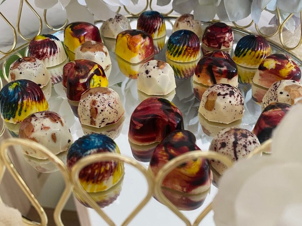 Elegantné ručne vyrábané čokoládové pralinky s umeleckým farebným dizajnom, servírované na zrkadlovom podnose – dokonalá lahôdka od LE BON cukrárskej výroby.